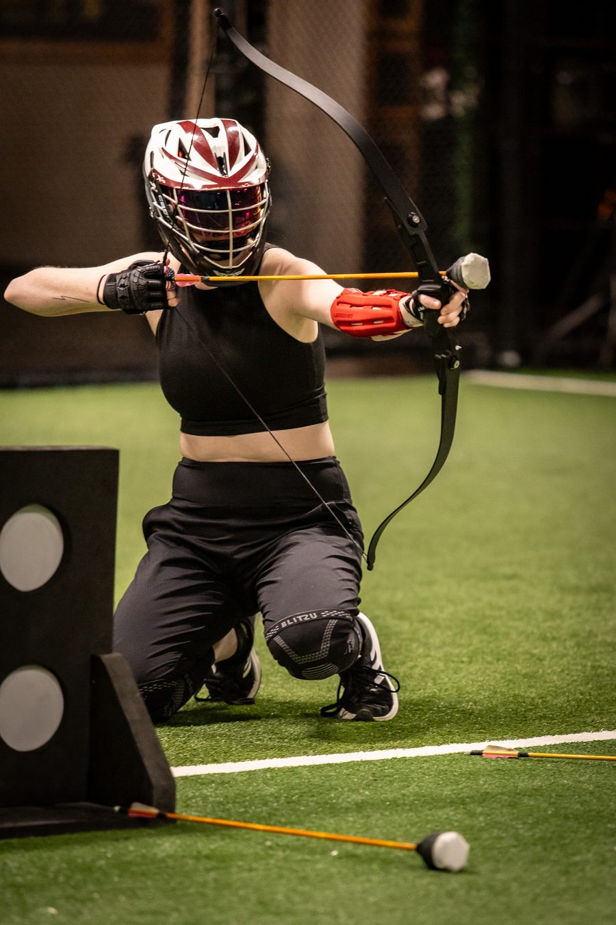 Trisha drawing a bow, kneeling, during a combat archery match